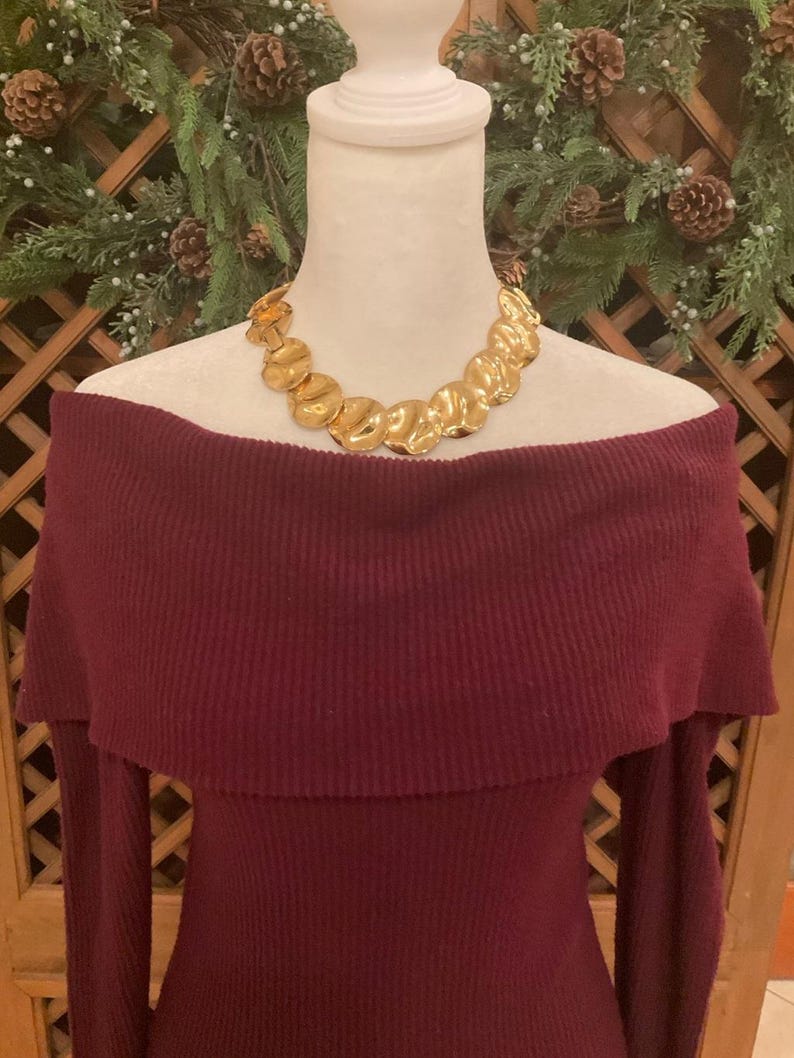 Puede incluir: Un vestido burdeos con hombros descubiertos y mangas largas se exhibe en un maniqu&iacute;. Un collar dorado con un dise&ntilde;o org&aacute;nico y texturizado adorna el escote. El fondo presenta un arreglo festivo de vegetaci&oacute;n y pi&ntilde;as.