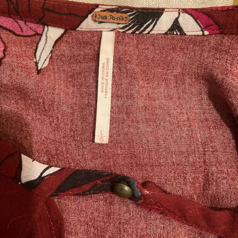 May include: Close-up of a burgundy-colored top with floral patterns in white, black, and pink. The tag reads "Free People" and "Made in China". The size is Large (L).