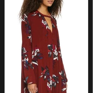 May include: A burgundy dress with a floral pattern. The dress has long sleeves, a keyhole neckline, and a loose fit. The floral design includes white, blue, and red accents. The dress is knee-length and made of a flowing fabric.