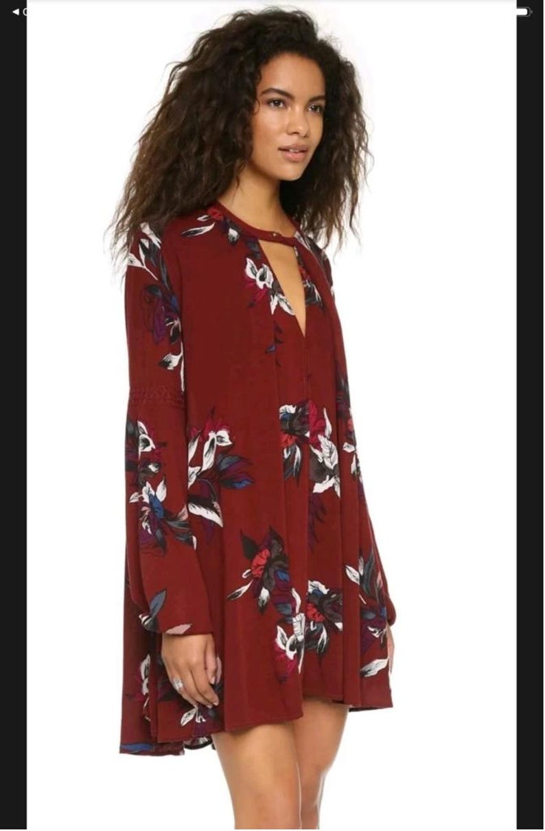 May include: A burgundy dress with a floral pattern. The dress has long sleeves, a keyhole neckline, and a loose fit. The floral design includes white, blue, and red accents. The dress is knee-length and made of a flowing fabric.