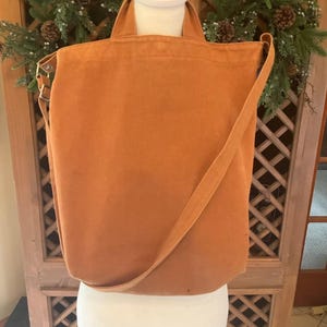 May include: A solid orange canvas tote bag featuring two top handles and a long adjustable shoulder strap. The bag has a rectangular shape with a flat base and is displayed on a white mannequin torso, showcasing its simple, functional design.