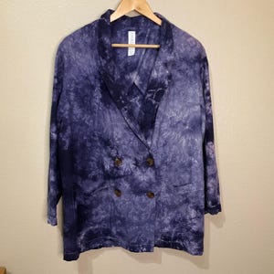 May include: A double-breasted blazer in a tie-dye pattern of blue and white. The jacket has a notched lapel, two front pockets, and brown buttons. The blazer is hanging on a wooden hanger.