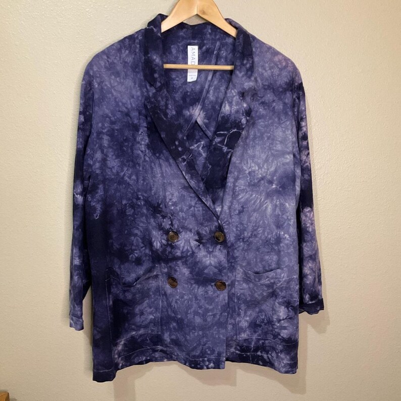 May include: A double-breasted blazer in a tie-dye pattern of blue and white. The jacket has a notched lapel, two front pockets, and brown buttons. The blazer is hanging on a wooden hanger.
