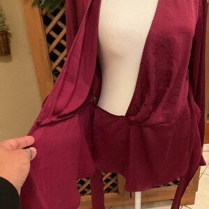 Puede incluir: Una blusa burdeos de manga larga con frente drapeado y cierre de lazo. La blusa est&aacute; hecha de un material sedoso y tiene un escote en V profundo. La prenda se exhibe en un maniqu&iacute;.