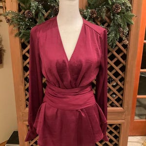 Puede incluir: Una blusa burdeos con mangas largas y cintura cruzada. La parte superior tiene un escote en V profundo y un dobladillo estilo peplum. La blusa se exhibe en un maniqu&iacute; blanco sobre un fondo de madera.