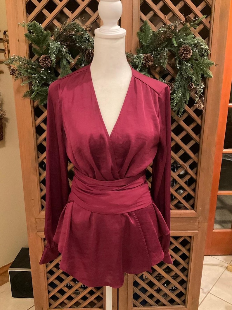 Puede incluir: Una blusa burdeos con mangas largas y cintura cruzada. La parte superior tiene un escote en V profundo y un dobladillo estilo peplum. La blusa se exhibe en un maniqu&iacute; blanco sobre un fondo de madera.