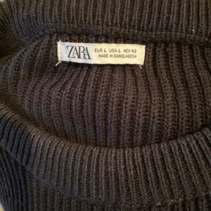 Pode incluir: Close-up de um su&eacute;ter de malha azul marinho escuro. O tecido de malha canelada &eacute; vis&iacute;vel, com uma etiqueta que diz "ZARA" e informa&ccedil;&otilde;es de tamanho. O su&eacute;ter parece ser de tamanho grande.