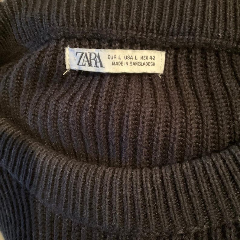 Pode incluir: Close-up de um su&eacute;ter de malha azul marinho escuro. O tecido de malha canelada &eacute; vis&iacute;vel, com uma etiqueta que diz "ZARA" e informa&ccedil;&otilde;es de tamanho. O su&eacute;ter parece ser de tamanho grande.