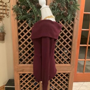 Puede incluir: Un su&eacute;ter burdeos con hombros descubiertos y mangas largas. El su&eacute;ter se exhibe en un maniqu&iacute; blanco. La tela de punto acanalado es de color rojo p&uacute;rpura intenso. Un collar dorado rodea el cuello del maniqu&iacute;.