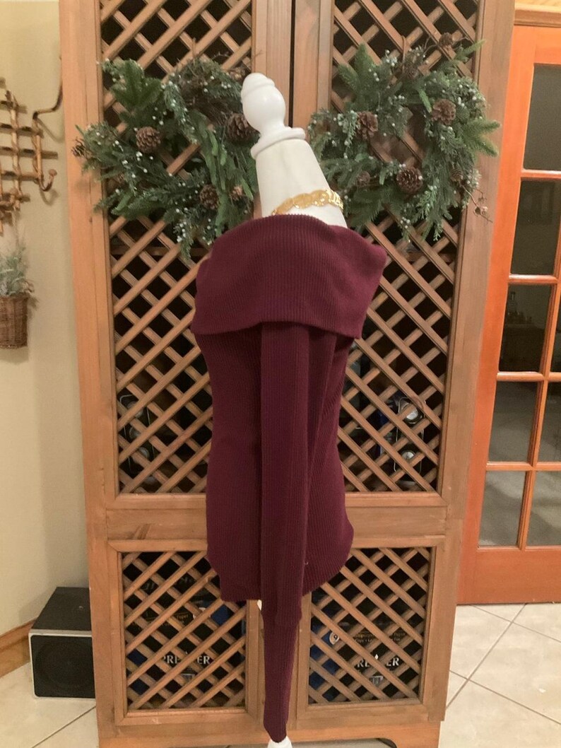 Puede incluir: Un su&eacute;ter burdeos con hombros descubiertos y mangas largas. El su&eacute;ter se exhibe en un maniqu&iacute; blanco. La tela de punto acanalado es de color rojo p&uacute;rpura intenso. Un collar dorado rodea el cuello del maniqu&iacute;.