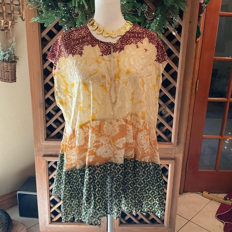 May include: A sleeveless, tiered dress on a mannequin. The top section is a maroon and cream floral print. The middle section is a yellow and orange floral print. The bottom section is a green geometric print. A gold necklace is around the mannequin's neck.