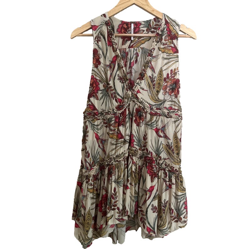 May include: A cream-coloured sleeveless top with a V-neck and a floral pattern in red, green, and gold. The top has a ruffled trim and is hanging on a wooden hanger.