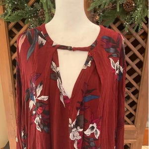 May include: A maroon long-sleeve top with a keyhole neckline and floral print. The top features a V-neck and a button closure at the neckline. The floral pattern includes white, pink, and blue accents.