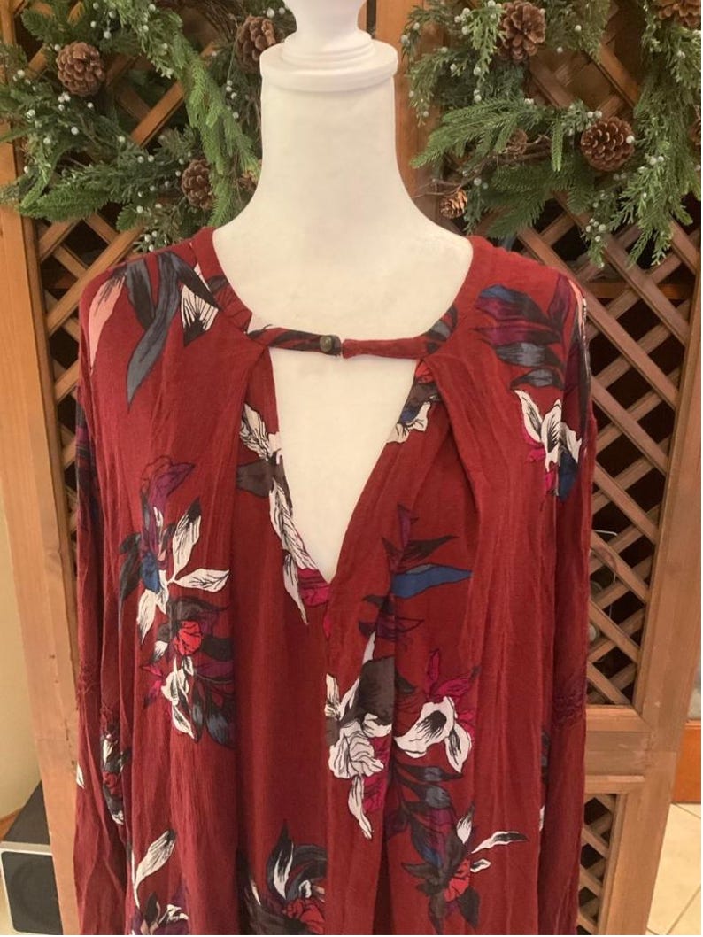 May include: A maroon long-sleeve top with a keyhole neckline and floral print. The top features a V-neck and a button closure at the neckline. The floral pattern includes white, pink, and blue accents.