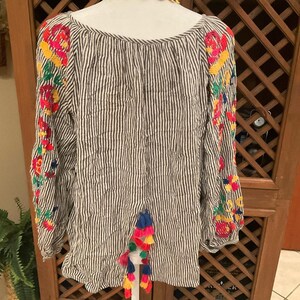 May include: A striped blouse with colourful floral embroidery on the sleeves. The long-sleeved top features a scoop neckline and a split back with vibrant tassel accents. The fabric has vertical black and white stripes.