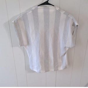 May include: A short-sleeved, striped shirt in white and light gray hangs on a black hanger. The vertical stripes run the length of the shirt. The shirt has a relaxed fit and a simple neckline. The fabric appears to be lightweight.