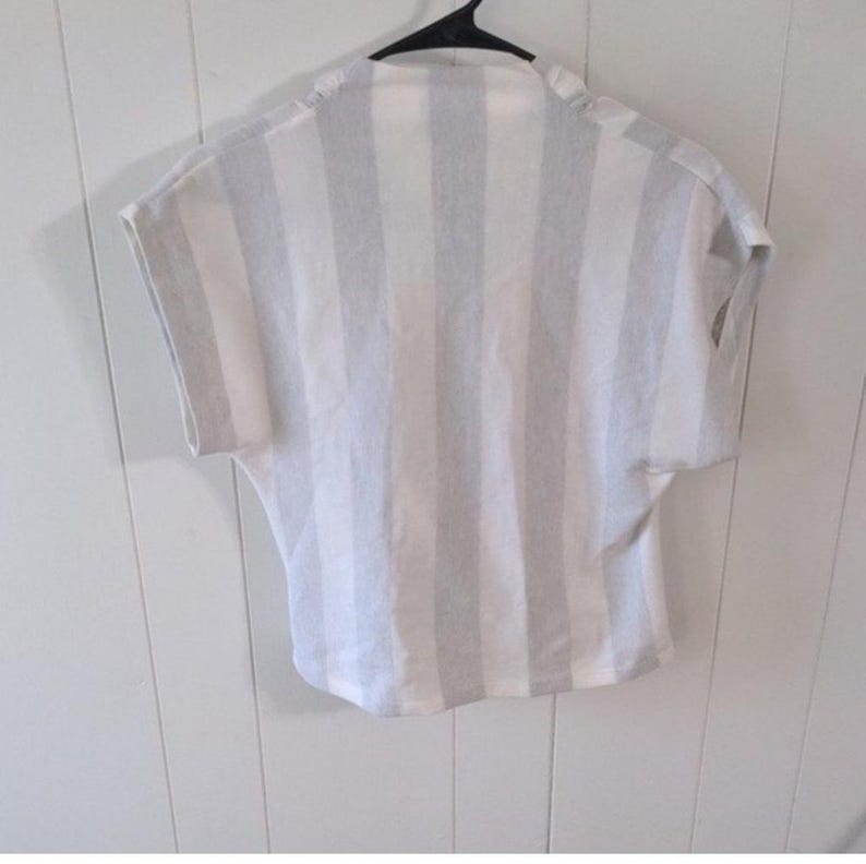 May include: A short-sleeved, striped shirt in white and light gray hangs on a black hanger. The vertical stripes run the length of the shirt. The shirt has a relaxed fit and a simple neckline. The fabric appears to be lightweight.