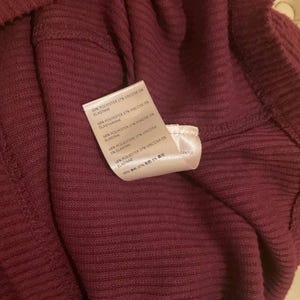 Puede incluir: Primer plano de una prenda burdeos con textura acanalada. Una etiqueta blanca muestra los detalles de la composici&oacute;n del tejido. La tela parece suave y tiene un ligero brillo. La prenda est&aacute; doblada, mostrando el interior y la textura.