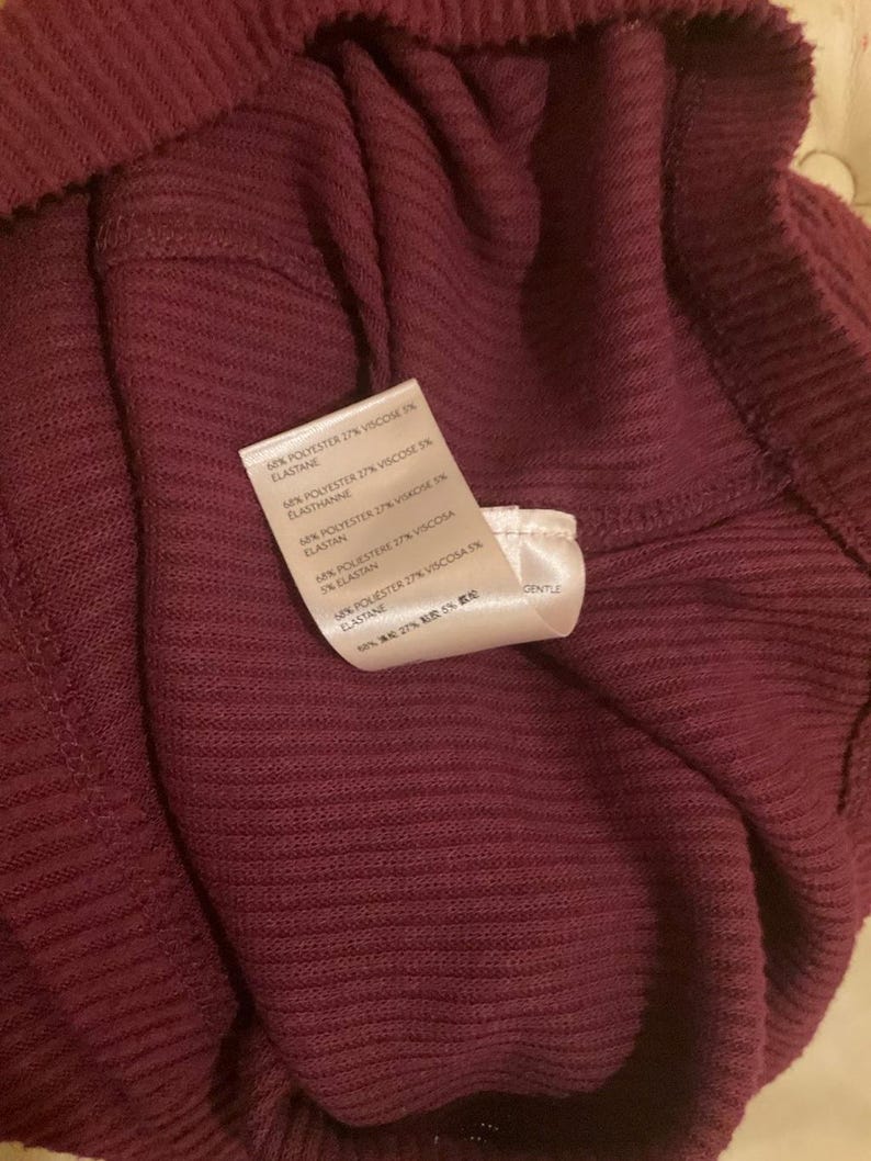 Puede incluir: Primer plano de una prenda burdeos con textura acanalada. Una etiqueta blanca muestra los detalles de la composici&oacute;n del tejido. La tela parece suave y tiene un ligero brillo. La prenda est&aacute; doblada, mostrando el interior y la textura.