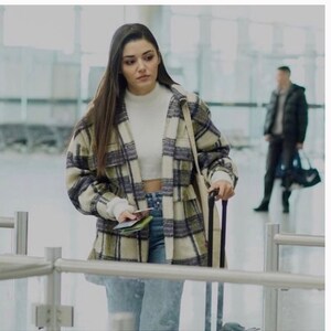 May include: A woman wearing a white shirt, blue jeans, and a plaid jacket is walking through an airport terminal. She is pulling a black suitcase behind her.