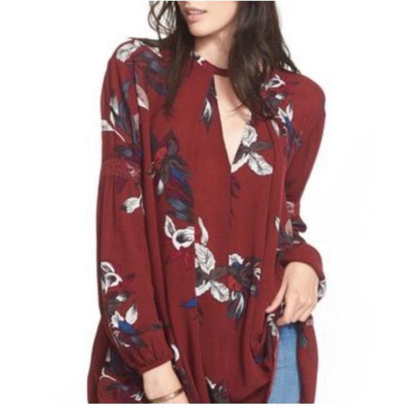 May include: A burgundy tunic top with a floral pattern. The long-sleeved top features a keyhole neckline and a relaxed fit. The floral design includes white, blue, and red accents, creating a bohemian style.