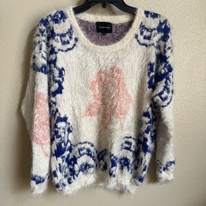 May include: White fuzzy sweater with a blue and pink geometric pattern. The sweater has a crew neck and long sleeves. The brand label says "La Fille Verte".