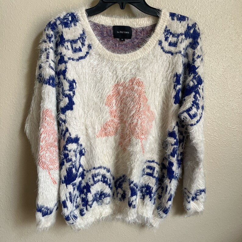May include: White fuzzy sweater with a blue and pink geometric pattern. The sweater has a crew neck and long sleeves. The brand label says "La Fille Verte".
