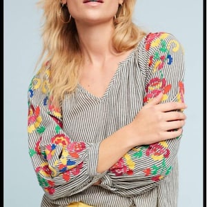 May include: A striped blouse with a V-neck and colourful floral embroidery on the sleeves. The blouse is paired with yellow trousers. The top features a black and white striped pattern.