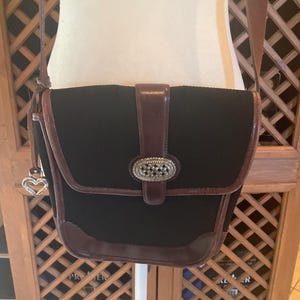 May include: Black crossbody bag with brown leather trim and a decorative silver-tone buckle. The bag features a flap closure and a long adjustable strap. A silver heart charm hangs from the side. The bag is displayed on a white stand.