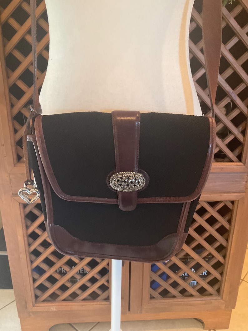 May include: Black crossbody bag with brown leather trim and a decorative silver-tone buckle. The bag features a flap closure and a long adjustable strap. A silver heart charm hangs from the side. The bag is displayed on a white stand.