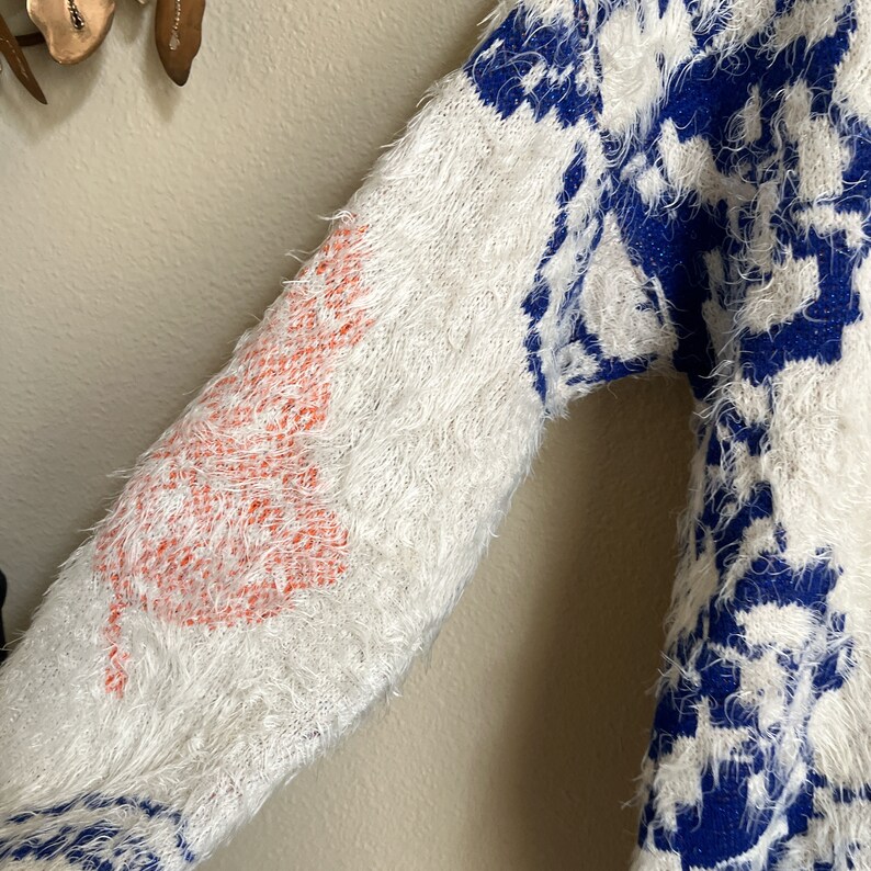 May include: A white fuzzy sweater with a blue and white patterned design. The sweater has a large orange tree design on the sleeve.