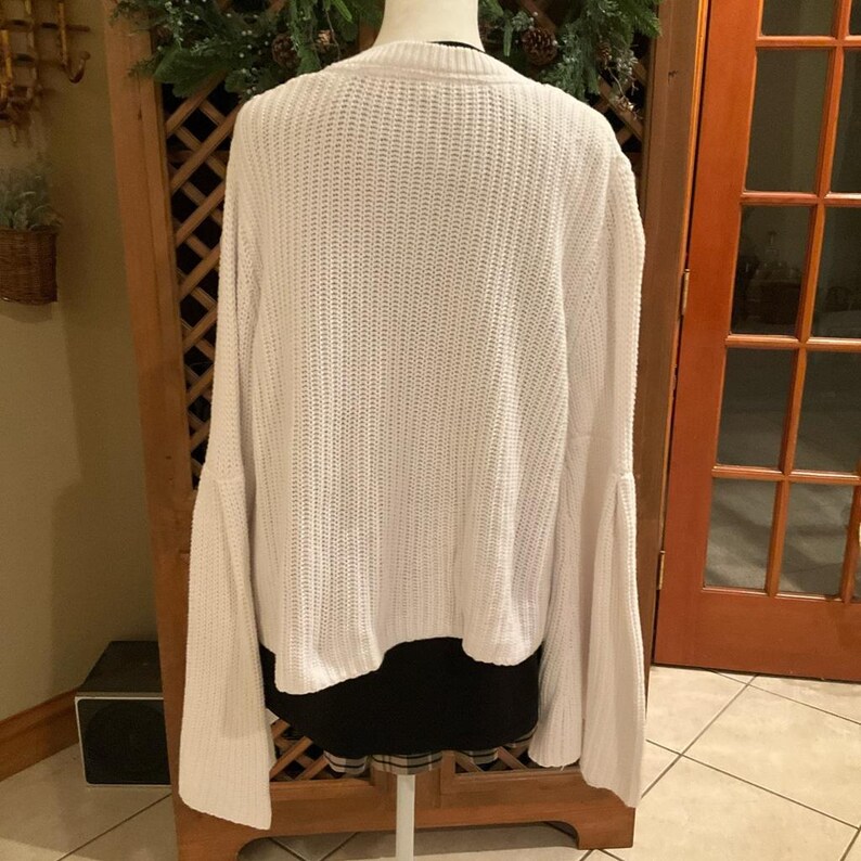May include: A white, long-sleeved sweater with bell sleeves. The sweater is made of a thick knit material and has a cropped length. The back of the sweater is visible, showing the texture of the knit.