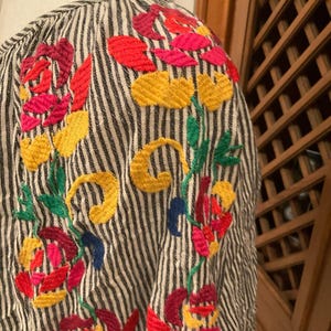 May include: Close-up of a striped shirt with colourful floral embroidery. The embroidery features red, pink, yellow, green, and blue thread, creating a vibrant design. The shirt's fabric has vertical black and white stripes.