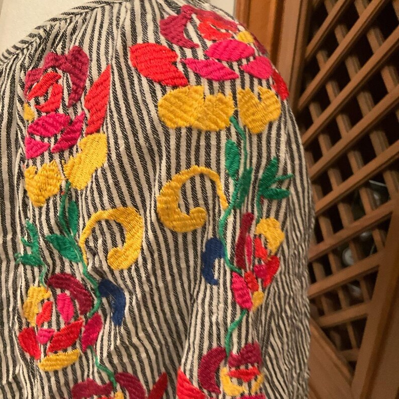 May include: Close-up of a striped shirt with colourful floral embroidery. The embroidery features red, pink, yellow, green, and blue thread, creating a vibrant design. The shirt's fabric has vertical black and white stripes.