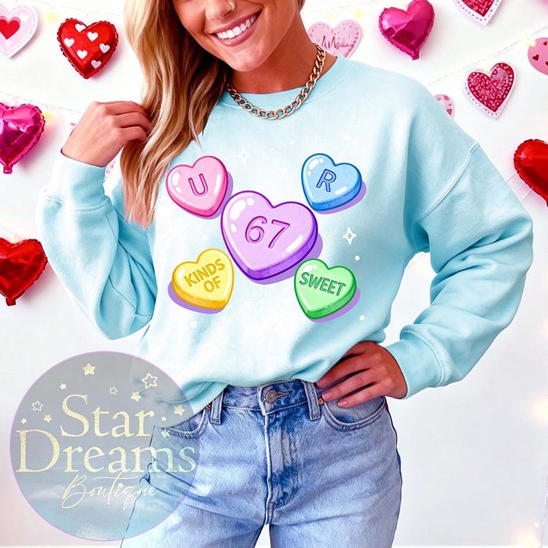 Op de afbeelding: Een lichtblauw sweatshirt met kleurrijke snoephartafbeeldingen. De harten spellen "U R 67 KINDS OF SWEET". De sweatshirt heeft een ronde hals en is gemaakt van een zacht materiaal. De afbeelding toont een persoon die de sweatshirt draagt.