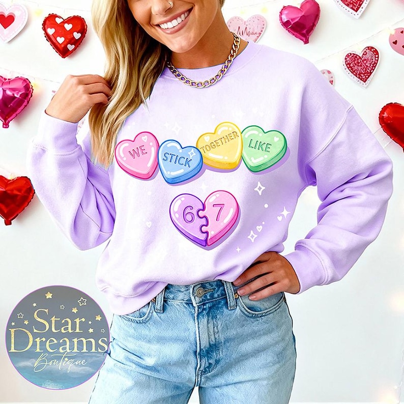 Pu&ograve; includere: Una felpa lilla con una grafica a forma di cuore con la scritta "We Stick Together Like" e un cuore a puzzle con i numeri "67". Il design ha un tema di San Valentino.