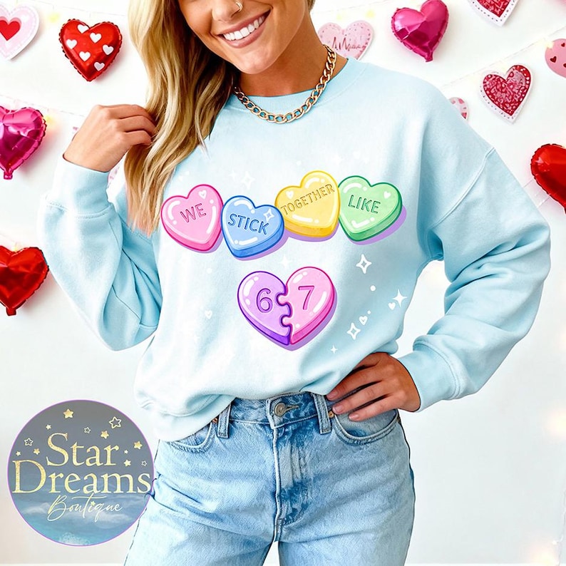 Pu&ograve; includere: Una felpa azzurra con un tema di San Valentino. Presenta caramelle a forma di cuore con le frasi "WE STICK TOGETHER LIKE" e un cuore a puzzle con i numeri "6 7". Il logo Star Dreams Boutique &egrave; visibile.