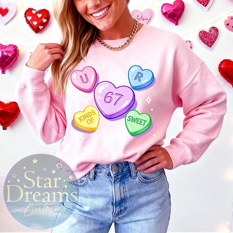 Op de afbeelding: Een lichtroze sweatshirt met kleurrijke snoephartafbeeldingen. De harten tonen de woorden "U", "R", "Sweet" en "Kinds of", samen met het nummer "67". Het sweatshirt maakt deel uit van de Star Dreams Boutique collectie.
