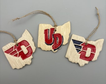 May include: Three ceramic ornaments shaped like the state of Ohio, each with a different University of Dayton logo. The logos are red and blue on a cream background.