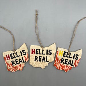 May include: Three wooden ornaments shaped like the state of Ohio, each with the text "Hell is Real" in black lettering with red and yellow accents. The ornaments are hanging from twine.