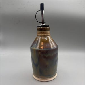 May include: A handcrafted ceramic oil bottle with a stainless steel spout and black cap. The bottle has a brown and blue glaze with a textured, ridged neck and a white base. The bottle is approximately 8 inches tall.