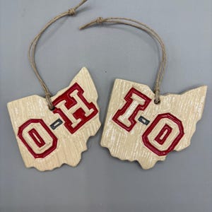 May include: Two ceramic ornaments shaped like the state of Ohio, each with the letters "O-H" in red on a white background. The ornaments are attached to twine.