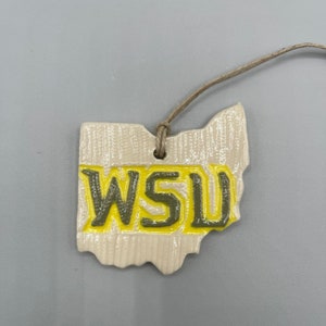 May include: A cream-colored ceramic ornament shaped like the state of Ohio, with the letters "WSU" in yellow and green. A brown string is attached for hanging. The ornament has a textured surface.