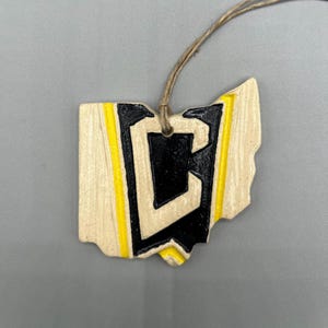 May include: A ceramic ornament shaped like the state of Ohio with a black letter "C" in the centre. The ornament is painted with yellow stripes and has a brown string for hanging.