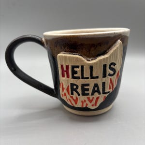 May include: A black ceramic mug with a brown glaze and a white design of the state of Ohio with the text "Hell is Real" in red and black lettering.