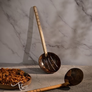 May include: Two wooden coconut shell ladles with brown handles. The ladles have a slotted design and are perfect for serving food. The ladles are on a white marble surface.