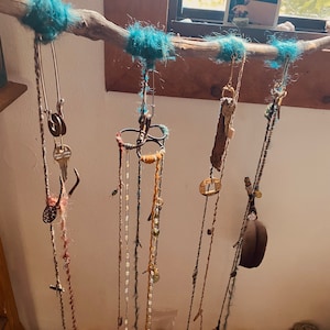 Puede incluir: Una rama de madera flotante con hilo turquesa envuelto alrededor, de la rama cuelgan varios hilos de lana con varios amuletos y baratijas adjuntos. Los amuletos incluyen llaves, cuentas, un pequeño anillo de metal y un pequeño sombrero marrón.