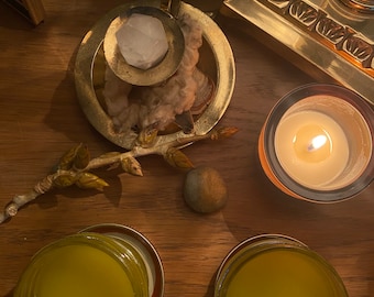 May include: A wooden table with two glass jars filled with a yellow liquid, a small candle in a glass jar, a small branch with leaves, a small stone, and a gold metal candle holder with a white crystal.