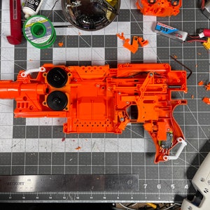 May include: An orange toy gun disassembled on a gray grid surface, revealing internal components. Various parts, including black circular pieces, white levers, and electronic components, are scattered around. Tools and batteries are also present, suggesting a repair or modification project.