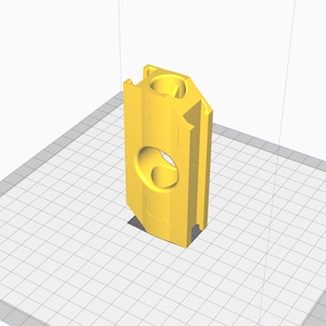 May include: A yellow 3D-printed object with a rectangular shape and a large, circular cutout. The object has a cylindrical top and is positioned on a grid-like surface. The design appears to be a component or part of a larger structure.
